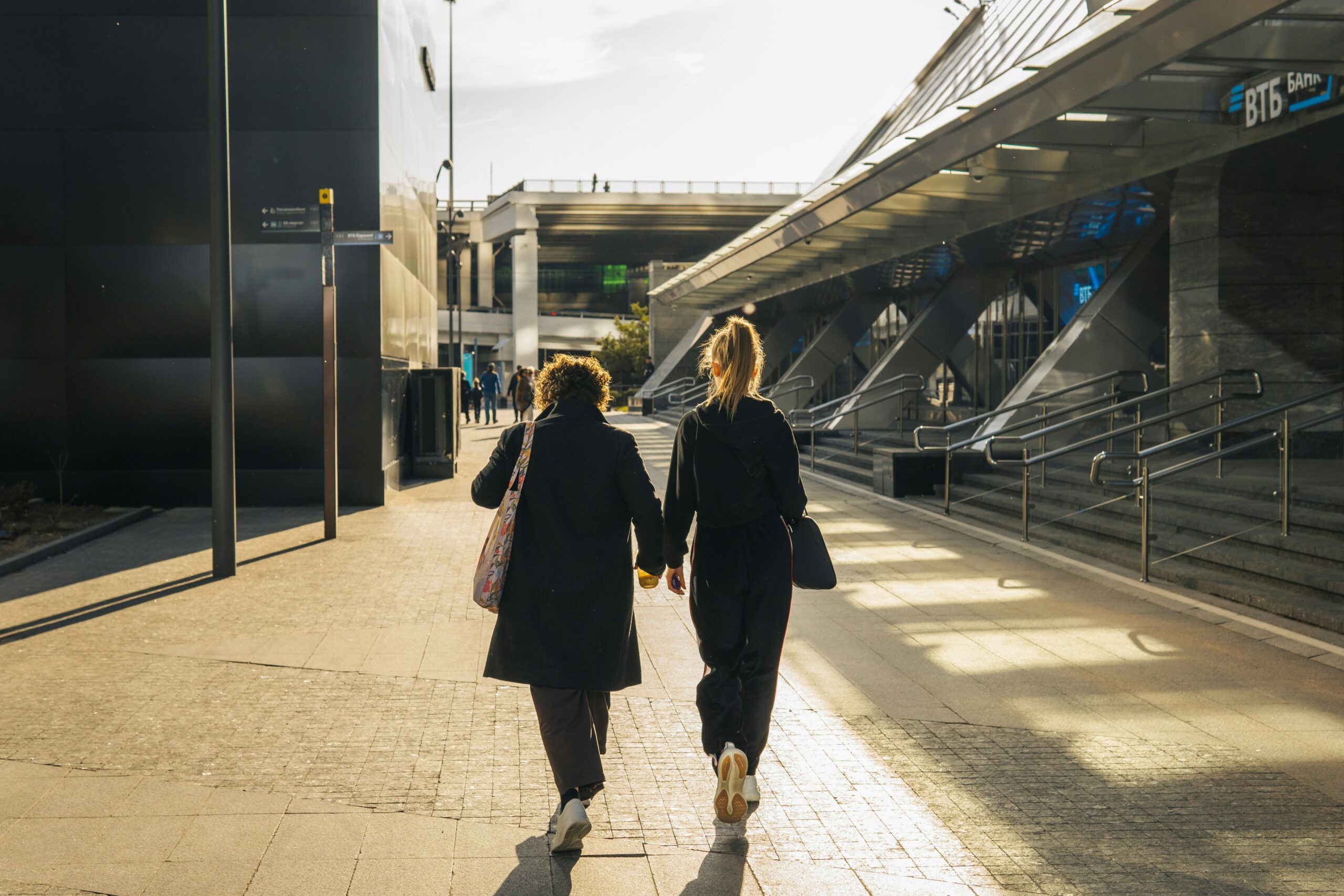 Two women walking side by side along a sunlit city walkway